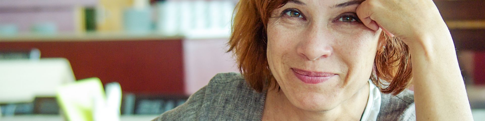Berufssprachkurse Lächelnde Frau mit roten Haaren sitzt am Tisch und schreibt mit einem orangen Stift in einem Heft