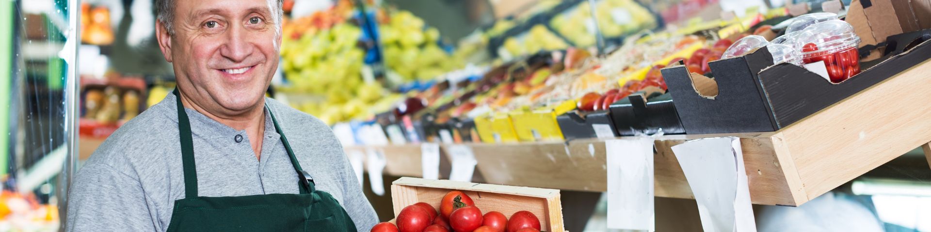 Quelle A | Sprachstart in die Arbeit Lächelnder Mann mit grünem Schürzen hält eine Kiste voller Tomaten in einem Obst- und Gemüseladen