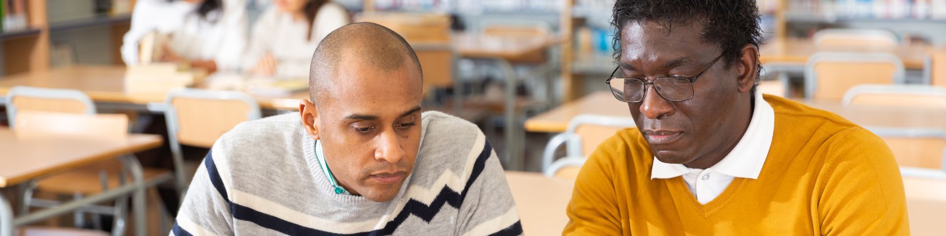 α-BIZ-Emsland Zwei Männer sitzen in einer Bibliothek und arbeiten gemeinsam an Notizen, umgeben von Büchern und Regalen.