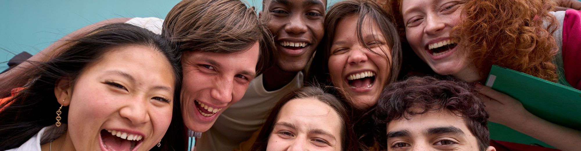 Gruppe von sieben jungen Menschen verschiedener Herkunft, die nah zusammen lachen und in die Kamera schauen