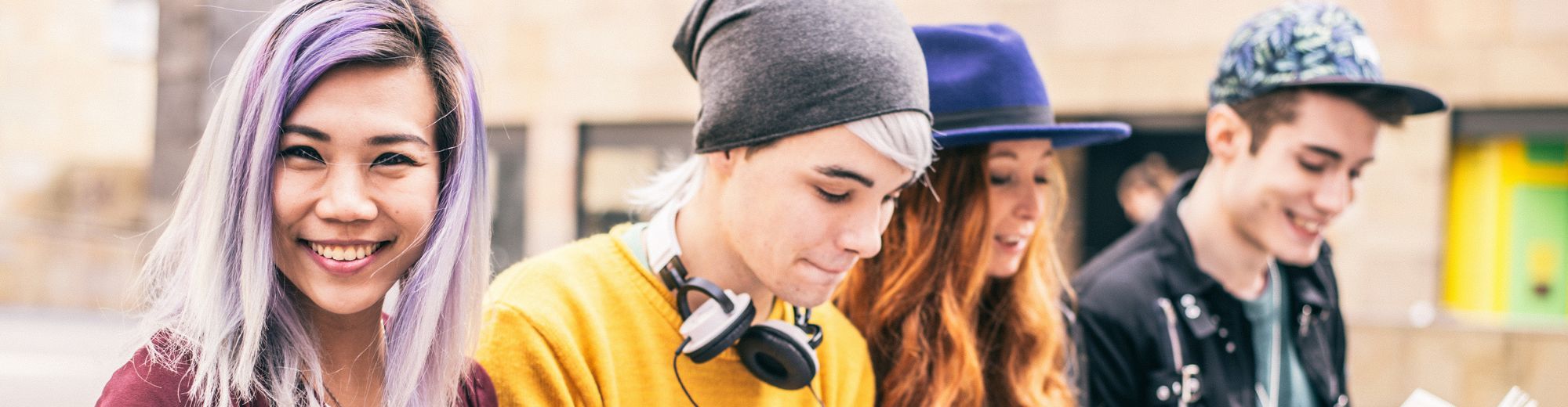 Vier junge Menschen sitzen nebeneinander, zwei lesen Bücher, einer benutzt ein Smartphone, einer hält ein Tablet, alle wirken entspannt und konzentriert