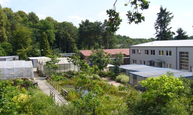 ZFJ Außenansicht eines Gebäudekomplexes mit Gewächshäusern und einem Teich, umgeben von Bäumen und Grünflächen